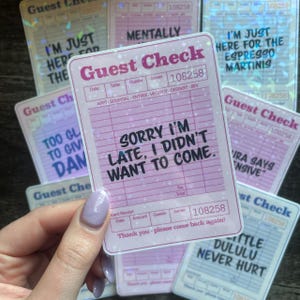 Peut inclure: Plusieurs autocollants roses et holographiques "Guest Check" avec des phrases amusantes. L'autocollant principal dit "Sorry I'm late, I didn't want to come." D'autres autocollants affichent des textes comme "I'm just here for the espresso martinis".