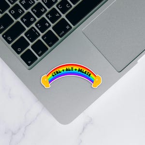Op de afbeelding: Een regenboogkleurige sticker in de vorm van een boog met gele handen aan beide uiteinden. De tekst CTRL + ALT + DELETE staat in zwarte hoofdletters in het midden van de regenboog gedrukt. De sticker is geplaatst op een zilveren laptop.