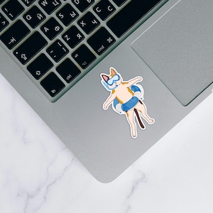 May include: A sticker featuring a cartoon cat wearing a blue snorkel and a blue and white inner tube. The cat is light brown and white with a dark brown tail. The sticker is placed on a silver laptop.