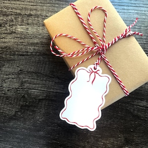 May include: A wrapped gift box in brown paper, tied with red and white twine. A white tag with a red border and bow hangs from the twine. The background is a dark wood surface.