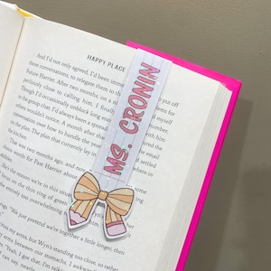 K&ouml;nnte beinhalten: Ein rosa-wei&szlig;es Lesezeichen mit Glitzer und einer Schleife. Das Lesezeichen tr&auml;gt den Text "MS. CRONIN" in rosa Buchstaben.