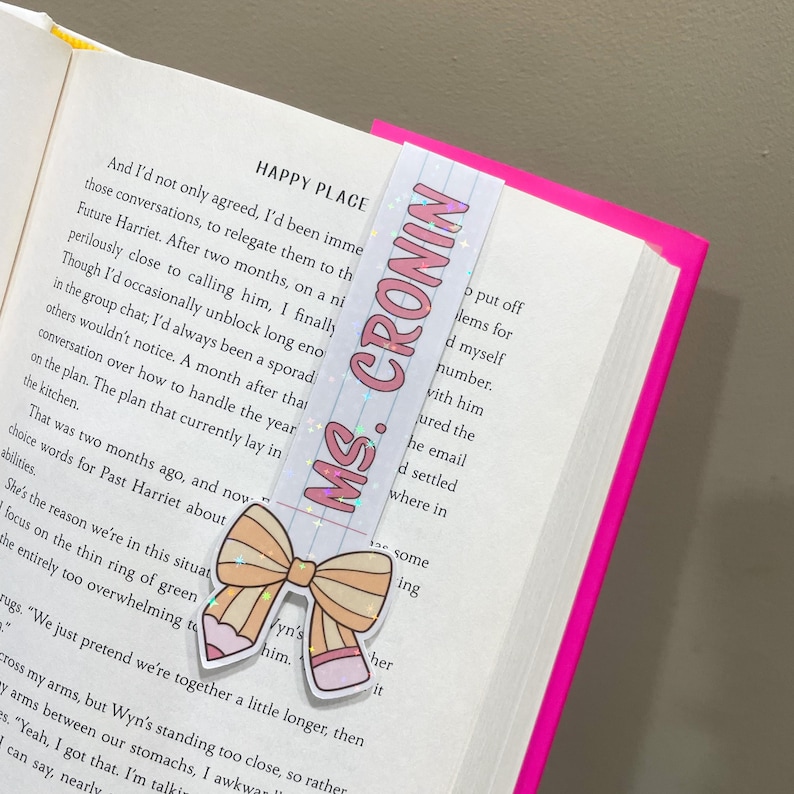 K&ouml;nnte beinhalten: Ein rosa-wei&szlig;es Lesezeichen mit Glitzer und einer Schleife. Das Lesezeichen tr&auml;gt den Text "MS. CRONIN" in rosa Buchstaben.