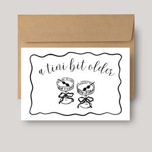 May include: A greeting card with a brown envelope. The white card has a black border and the text "a tini bit older". Two martini glasses with olives are illustrated below the text, each with a bow.