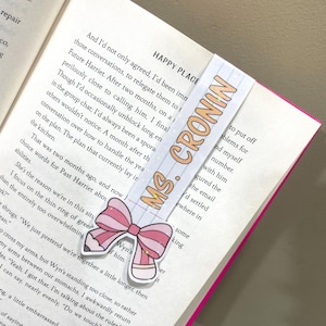 K&ouml;nnte beinhalten: Ein pinkfarbenes und wei&szlig;es Schleifen-Lesezeichen mit Glitzerakzenten. Das Lesezeichen tr&auml;gt den Text "MS. CRONIN" in orangefarbenen Buchstaben. Das Lesezeichen befindet sich auf einem Buch mit dem Text "HAPPY PLACE" auf dem Cover.