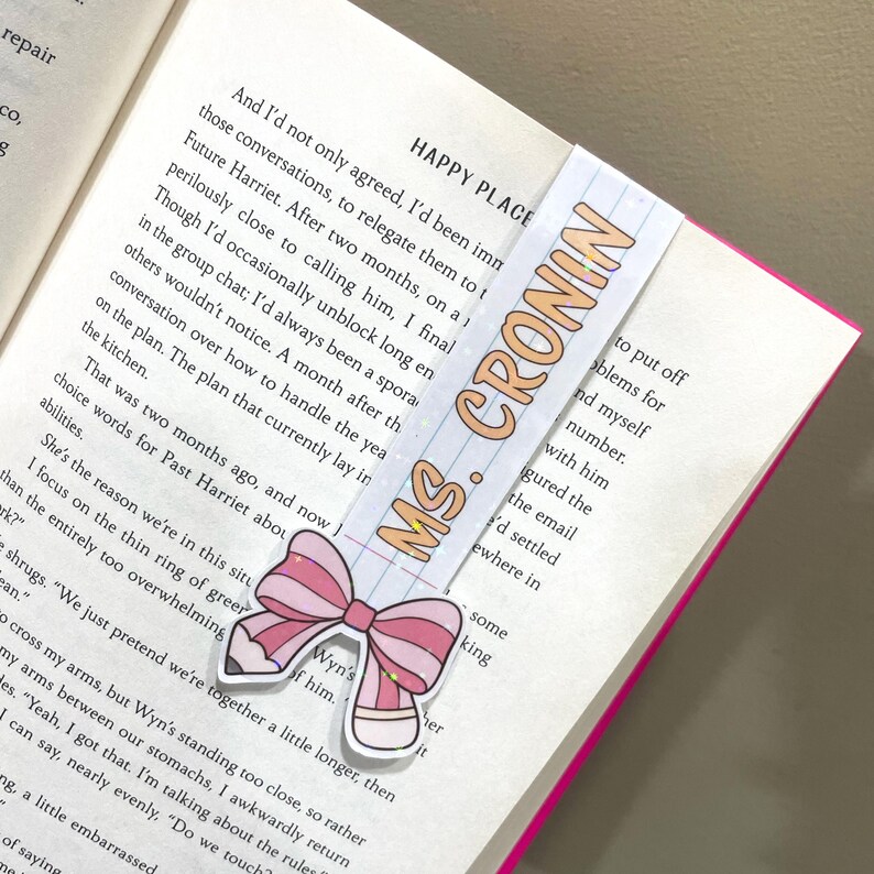 K&ouml;nnte beinhalten: Ein pinkfarbenes und wei&szlig;es Schleifen-Lesezeichen mit Glitzerakzenten. Das Lesezeichen tr&auml;gt den Text "MS. CRONIN" in orangefarbenen Buchstaben. Das Lesezeichen befindet sich auf einem Buch mit dem Text "HAPPY PLACE" auf dem Cover.