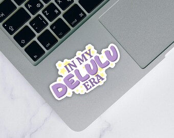 May include: A sticker with the text "IN MY DELULU ERA" in purple bubble letters, outlined in white, with yellow star accents. The sticker is on a silver laptop.