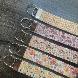 May include: Five fabric key fobs with silver-coloured hardware. The key fobs feature autumn-themed patterns with leaves, pumpkins, and mushrooms in shades of orange, yellow, and burgundy. The key fobs are arranged diagonally.