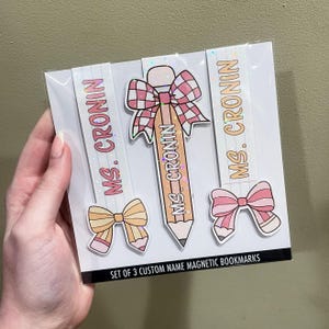 K&ouml;nnte beinhalten: Set mit drei benutzerdefinierten magnetischen Lesezeichen mit einem rosa und wei&szlig;en Bleistift, einer rosa und wei&szlig;en Schleife und einer rosa und wei&szlig;en Schleife mit einem Bleistift. Die Lesezeichen sind mit Glitzer verziert und der Text "MS. CRONIN" ist auf jedem Lesezeichen gedruckt.