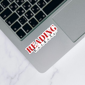 May include: A white sticker with the words "READING Rockets" in red and black script is placed on a silver laptop. The laptop is open, and the keyboard is visible. The sticker is rectangular and has a white border.