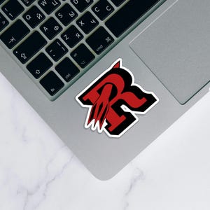 May include: A sticker featuring a stylized red letter "R" with a black outline and a red flame-like design. The sticker is placed on a silver laptop, with a black keyboard visible.