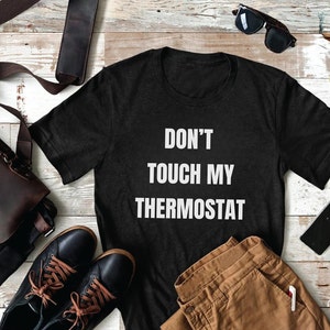 May include: A black t-shirt with the text "Don't Touch My Thermostat" in white lettering. The shirt is laid out on a wooden surface with a brown leather bag, sunglasses, and brown pants.