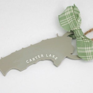 May include: A gray, map-shaped ornament with the text "CARTER LAKE" in white. It has a green and white plaid ribbon and a twine loop for hanging. The ornament is likely a souvenir or decorative item.