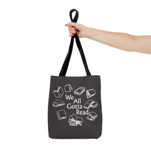 May include: A black tote bag with two black handles featuring a white graphic print of various open and closed books arranged in a circle around the centered white text that reads We All Gotta Read in a clean, serif font style.