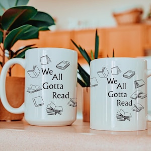 May include: Two white ceramic mugs featuring a black printed design of various books scattered around the text "We All Gotta Read". The mugs have a cylindrical shape with a curved handle on the side, displaying the same graphic and text on each.