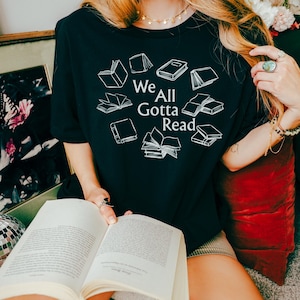 May include: A black t-shirt featuring white line art illustrations of various open and closed books arranged in a circular pattern around the text "We All Gotta Read" printed in the center in a clean, white, sans-serif font.