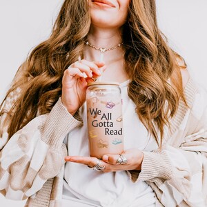 May include: A clear glass tumbler with a bamboo lid and a straw, filled with a light brown beverage. The glass features the black text "We All Gotta Read" surrounded by small, colorful illustrations of open and closed books in various colors.