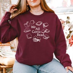 May include: A maroon long-sleeve crewneck sweatshirt featuring a white graphic print of various open and closed books arranged in a circle around the text "We All Gotta Read" written in a clean, white, sans-serif font centered on the chest.