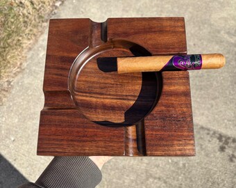 May include: A dark brown wooden cigar ashtray with a round indentation for the cigar and a smaller indentation for ashes. A brown cigar with a purple and gold band is resting in the ashtray.