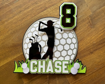 Peut inclure: Un topper de gâteau sur le thème du golf, de couleur verte, blanche et noire, avec une silhouette de golfeur, une balle de golf et le chiffre 8. Le nom "CHASE" est écrit en lettres blanches sur un fond vert.