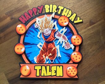 以下が含まれることがあります： ドラゴンボールZのキャラクターをあしらった、多層構造のケーキトッパー。「HAPPY BIRTHDAY TALEN」の文字が黄色と赤でデザインされています。キャラクターはオレンジと青で描かれ、オレンジ色の球体で囲まれています。