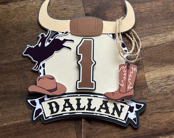 Könnte beinhalten: Ein brauner und weißer Cowboy-Kuchentopper mit einem braunen Bullenkopf, einem braunen Cowboyhut, einem braunen Cowboystiefel, einer schwarzen Silhouette eines Cowboys, der einen Bullen reitet, und der Zahl 1 in Braun. Der Topper enthält auch ein Banner mit dem Namen "Dallan" in Schwarz.