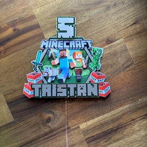 May include: A Minecraft themed cake topper featuring the number 5, the word "Minecraft", and the name "Tristan" in green and black lettering. The topper includes characters from the game, such as Steve and Alex, and TNT blocks.