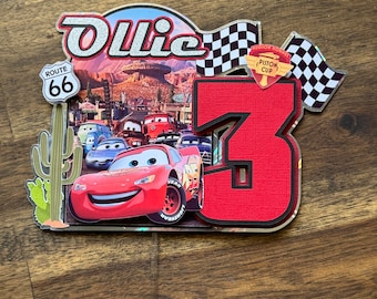 Puede incluir: Un adorno para pastel de Cars de color rojo, blanco y negro con el número 3, una bandera a cuadros y las palabras "Ollie" y "Piston Cup".