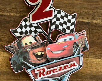 Puede incluir: Un adorno para pastel de bandera a cuadros roja y negra con un número 2 rojo, un coche de carreras rojo y una grúa roja. El adorno para pastel también tiene la palabra "Roczen" en letras rojas de purpurina.