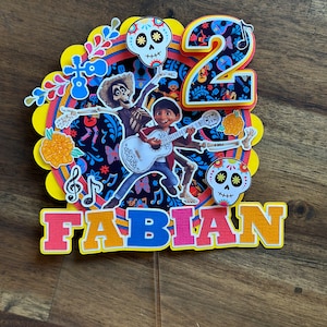 May include: A colourful cake topper featuring characters from the film Coco. The topper includes a skeleton playing a guitar, a boy with a guitar, a skull with flowers, and the number 2. The topper also includes the name "FABIAN" in colourful letters.