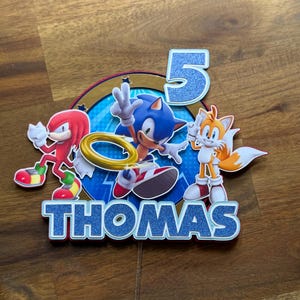 May include: A blue, red and yellow Sonic the Hedgehog cake topper featuring the characters Sonic, Tails and Knuckles. The topper also includes the number 5 and the name Thomas.