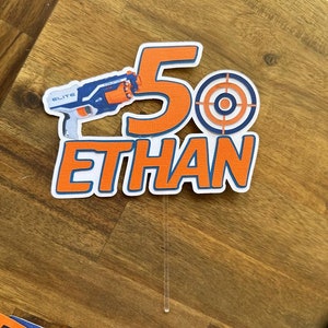 May include: Orange and blue cake topper featuring the number 5, the name Ethan, a target, and a toy gun. The gun is blue and orange and has the word "Elite" on it.