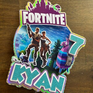 以下が含まれることがあります： Fortniteをテーマにしたケーキトッパー。紫と白の「Fortnite」ロゴ、青と紫のラマ、2人のキャラクター、そしてキラキラの仕上げが施された青い「Kyan」という名前が書かれています。