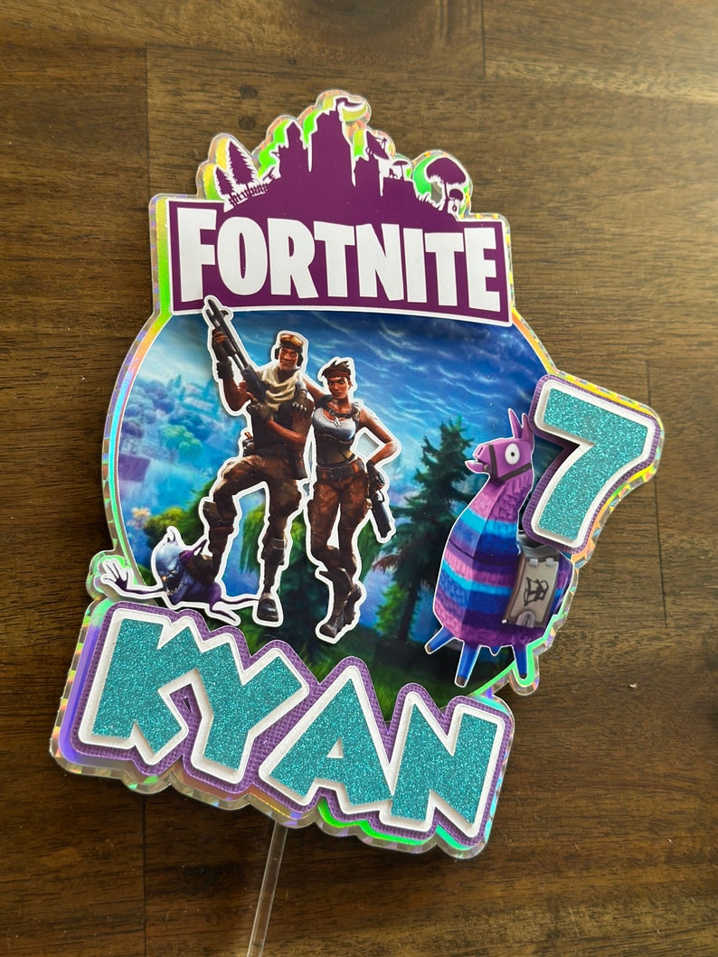 以下が含まれることがあります： Fortniteをテーマにしたケーキトッパー。紫と白の「Fortnite」ロゴ、青と紫のラマ、2人のキャラクター、そしてキラキラの仕上げが施された青い「Kyan」という名前が書かれています。