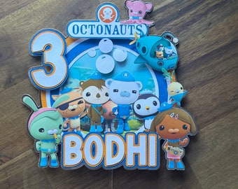 May include: A blue and orange cake topper featuring the characters from the cartoon show Octonauts. The topper says "Octonauts" and "Bodhi" and includes the number "3".