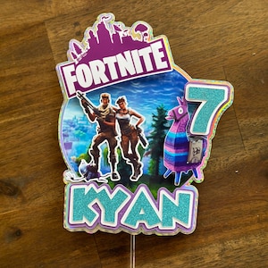 以下が含まれることがあります： ラマ、2人のキャラクター、そして「Fortnite」と「Kyan 7」の文字が書かれた、紫と青のフォートナイトのケーキトッパーです。