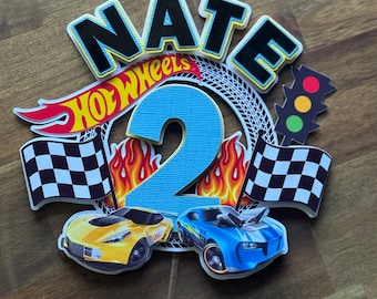 May include: A cake topper featuring the name "NATE" in black letters with a yellow outline, a blue number "2", a red, yellow, and black Hot Wheels logo, two checkered flags, two toy cars, and a traffic light.