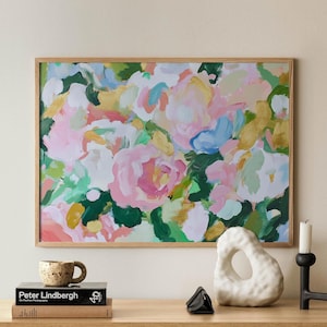 May include: A framed abstract painting with a floral theme. The painting features pink, white, green, and gold colors. The painting is hanging on a wall above a wooden cabinet with a rattan front.