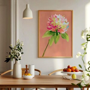 May include: A framed print of a pink, yellow, and green flower with a peach background. The print is hanging on a white wall above a wooden table with a white vase and a bowl of fruit.
