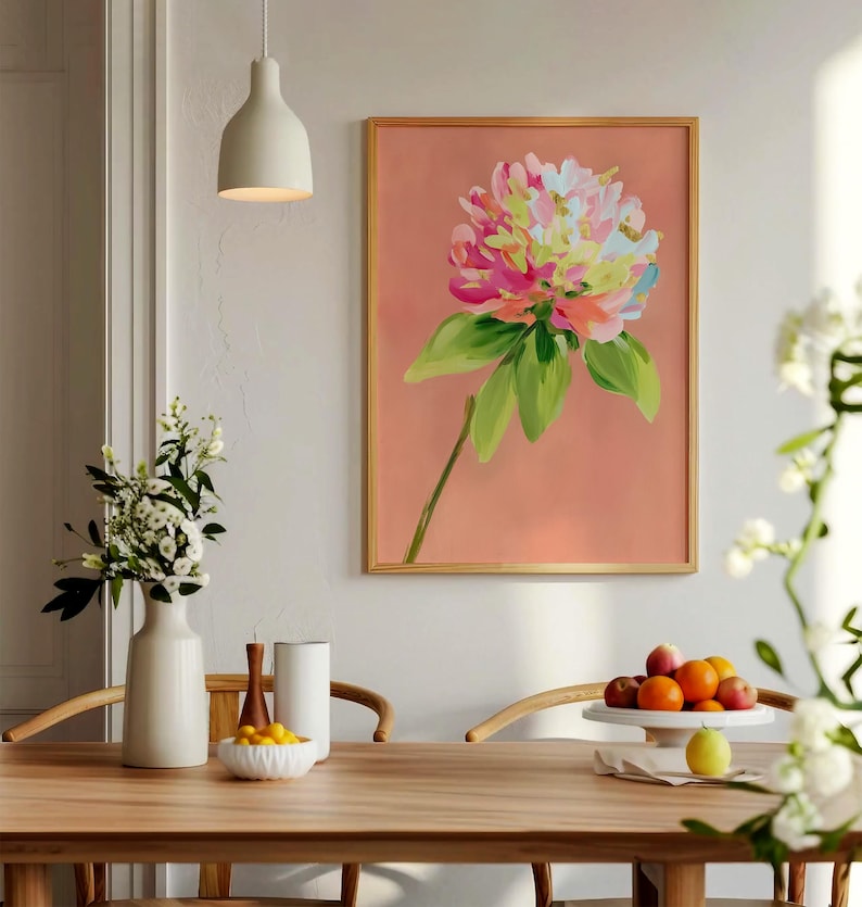 May include: A framed print of a pink, yellow, and green flower with a peach background. The print is hanging on a white wall above a wooden table with a white vase and a bowl of fruit.
