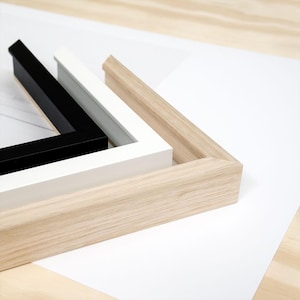 K&ouml;nnte beinhalten: Drei Bilderrahmen in Schwarz, Wei&szlig; und Naturholzt&ouml;nen sind auf einer wei&szlig;en Oberfl&auml;che angeordnet. Die Rahmen sind rechteckig mit abgeschr&auml;gter Kante und zeigen ein modernes Design.