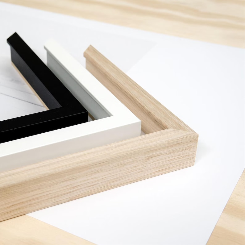 K&ouml;nnte beinhalten: Drei Bilderrahmen in Schwarz, Wei&szlig; und Naturholzt&ouml;nen sind auf einer wei&szlig;en Oberfl&auml;che angeordnet. Die Rahmen sind rechteckig mit abgeschr&auml;gter Kante und zeigen ein modernes Design.