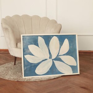 K&ouml;nnte beinhalten: Gerahmtes abstraktes Gem&auml;lde mit einer gro&szlig;en wei&szlig;en Blume vor blauem Hintergrund. Das Kunstwerk befindet sich in einem wei&szlig;en Rahmen und liegt auf einem grauen Teppich. Das Gem&auml;lde steht in der N&auml;he eines beigen Sessels.