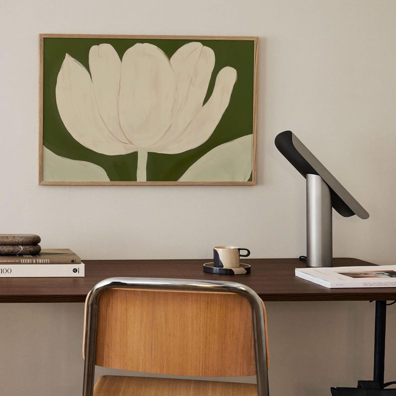 Peut inclure: Une impression encadr&eacute;e d'une fleur blanche avec des feuilles vertes sur un fond vert. L'impression est accroch&eacute;e &agrave; un mur blanc au-dessus d'un bureau avec une lampe, une tasse et une soucoupe, et un livre.