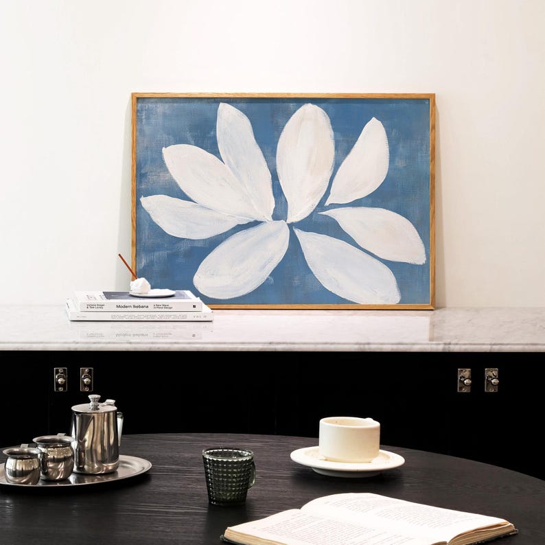 K&ouml;nnte beinhalten: Gerahmter Kunstdruck mit einer gro&szlig;en wei&szlig;en Blume vor blauem Hintergrund. Das Kunstwerk ist in einem Naturholzrahmen. B&uuml;cher und ein kleiner R&auml;ucherst&auml;bchenhalter liegen auf einer Marmoroberfl&auml;che.