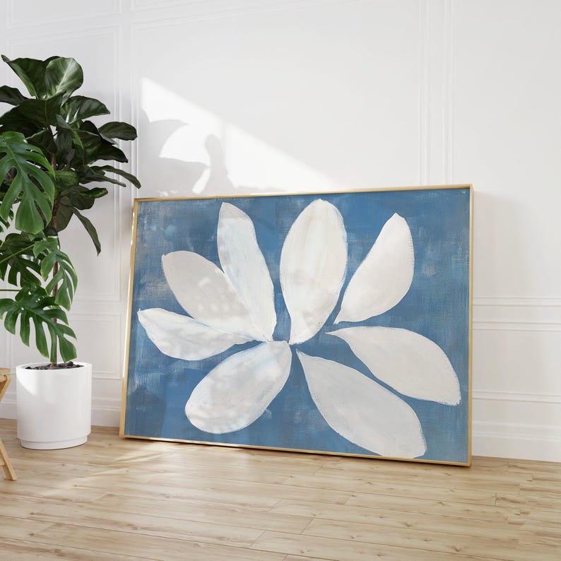 May include: A framed abstract painting with a blue background and white flower-like shapes. The painting is in a gold frame and is hanging on a white wall.