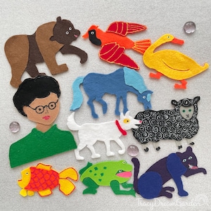 Puede incluir: Una colección de figuras de animales de fieltro de colores, que incluyen un oso, un pájaro, un pato, un caballo, un perro, una oveja, un gato, un pez y una rana. También se incluye una figura de persona con gafas. Las figuras están dispuestas sobre una superficie gris claro.