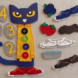May include: A felt cat character with a yellow shirt and red shoes, surrounded by felt objects including numbers, shoes, berries, and a bowl. The cat has a blue body, yellow eyes, and a white nose.