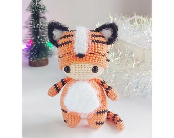 May include: A crocheted tiger plush toy wearing an orange and black striped hat with white fur accents. The toy has black eyes and a white face.