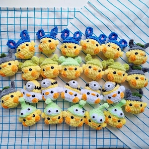 Könnte beinhalten: Eine Sammlung handgefertigter Häkelküken-Spielzeuge in verschiedenen Stilen. Die Küken sind hauptsächlich gelb mit Akzenten in Blau, Grün, Grau und Weiß. Einige haben Hasenohren, andere Hüte. Die Spielzeuge sind auf einer blau-weiß karierten Oberfläche angeordnet.