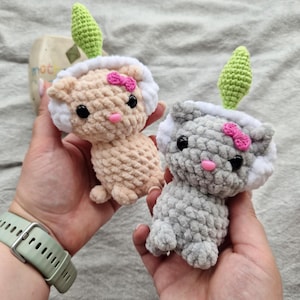 May include: Two crocheted cat toys, one beige and one grey, each with white trim, pink bows, and green leaf accents. The toys have black eyes and pink noses. The toys are held in hands.